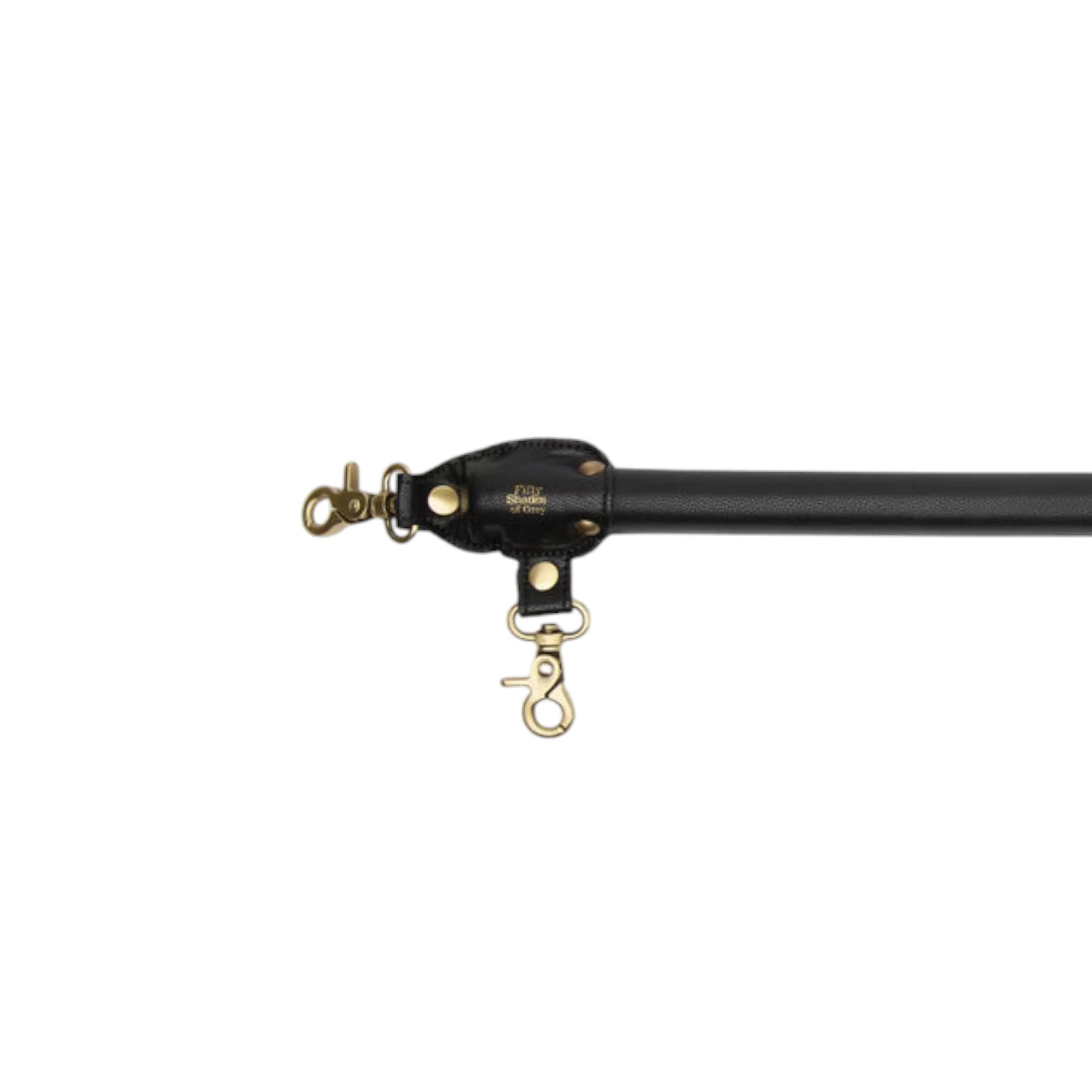 Fifty Shades of Grey Bound to You faux leather spreader bar shown close up displaying black faux leather end cap with gold rivets, gold-toned swivel clip and D-ring attachment points, and textured black bar against white background