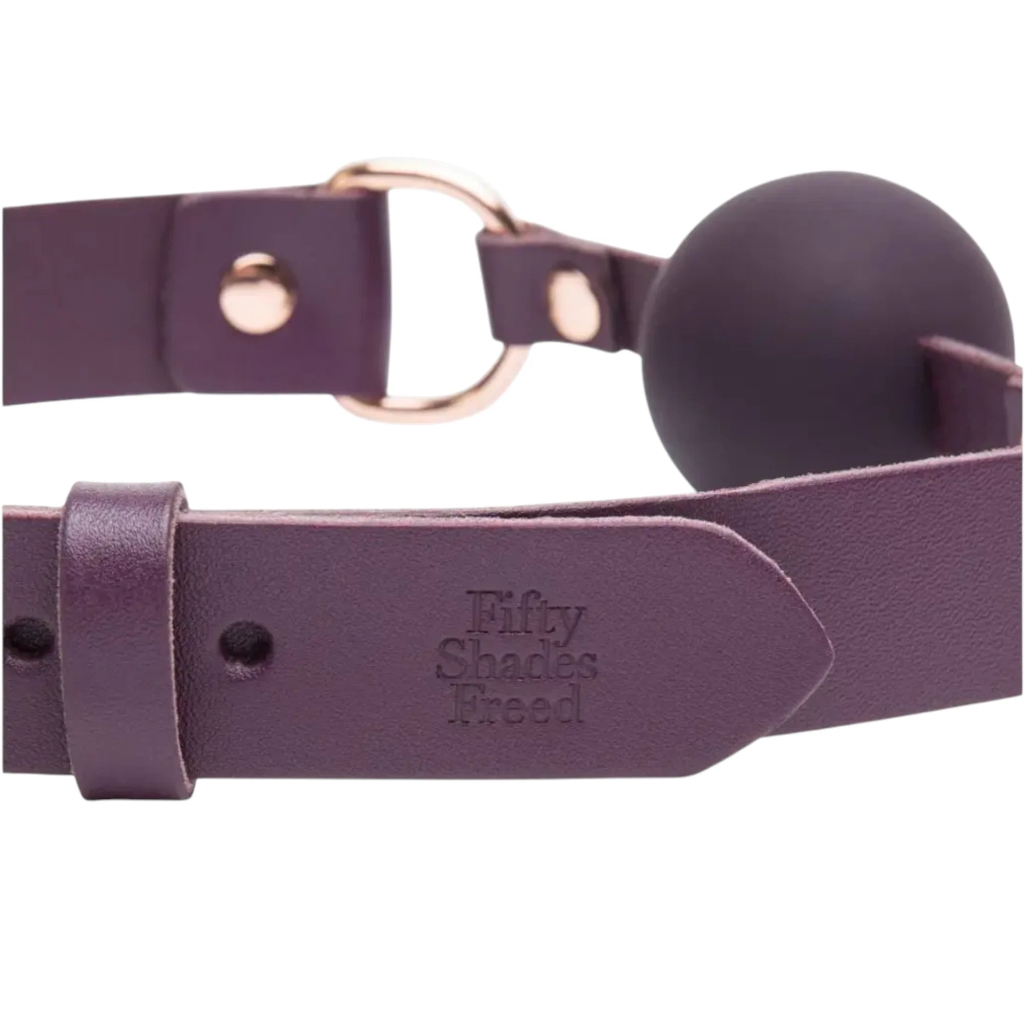 Close-up of the Fifty Shades Freed Cherished Collection leather ball gag, showing the purple strap with embossed Fifty Shades Freed branding, metal ring hardware and the dark silicone ball at the centre against a white background.