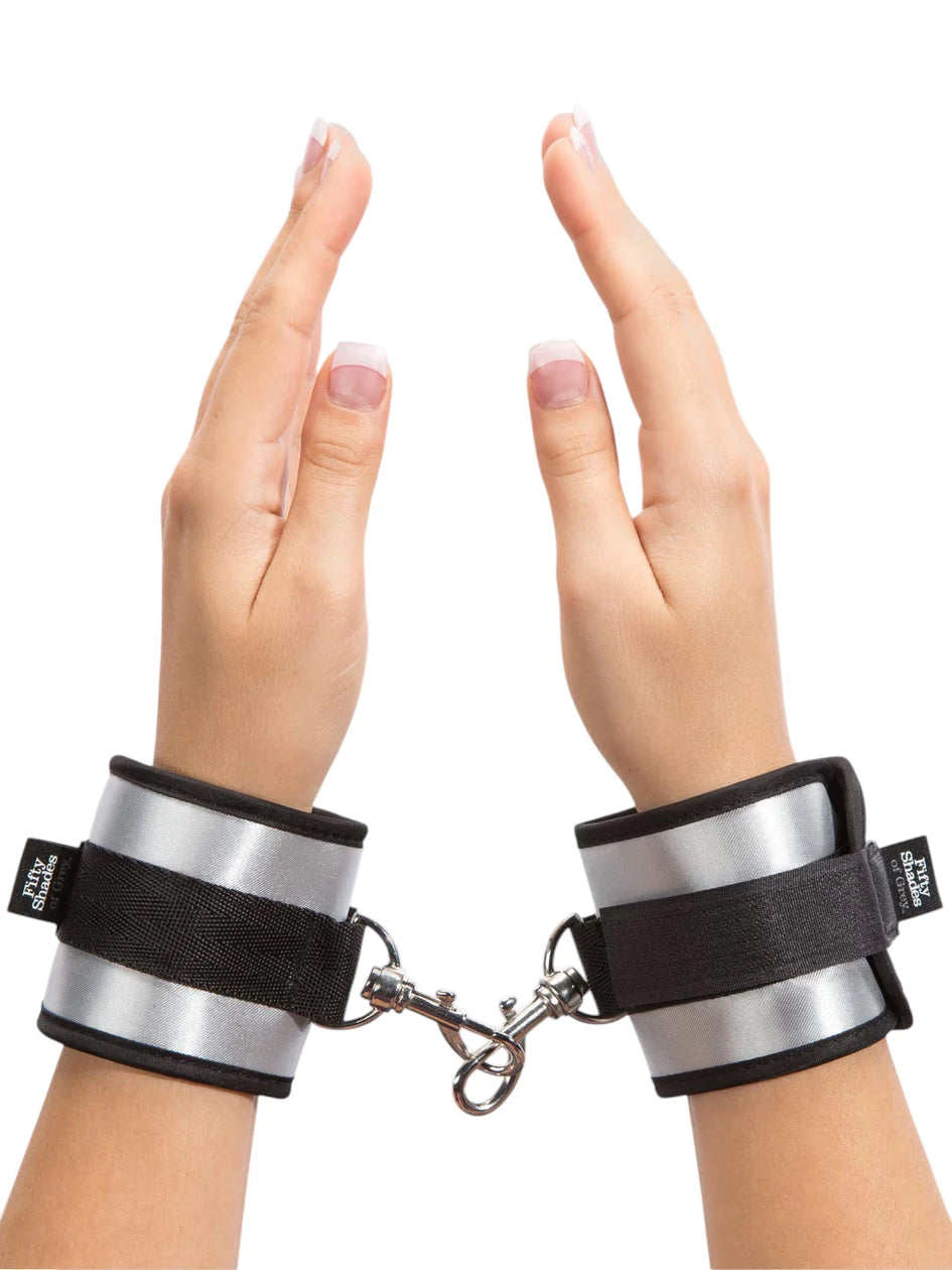 Fifty Shades of Grey Totally His soft handcuffs worn on wrists showing black neoprene lining, silver metallic exterior panels, black velcro fastening straps with Fifty Shades of Grey branding, and metal chain connector with D-rings against white background