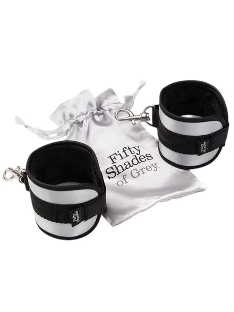 Fifty Shades of Grey Totally His soft handcuffs displayed with white satin storage pouch featuring drawstring closure and 'Fifty Shades of Grey' branding, showing connected pair with black neoprene lining, silver metallic panels, velcro straps, and metal chain connector against white background