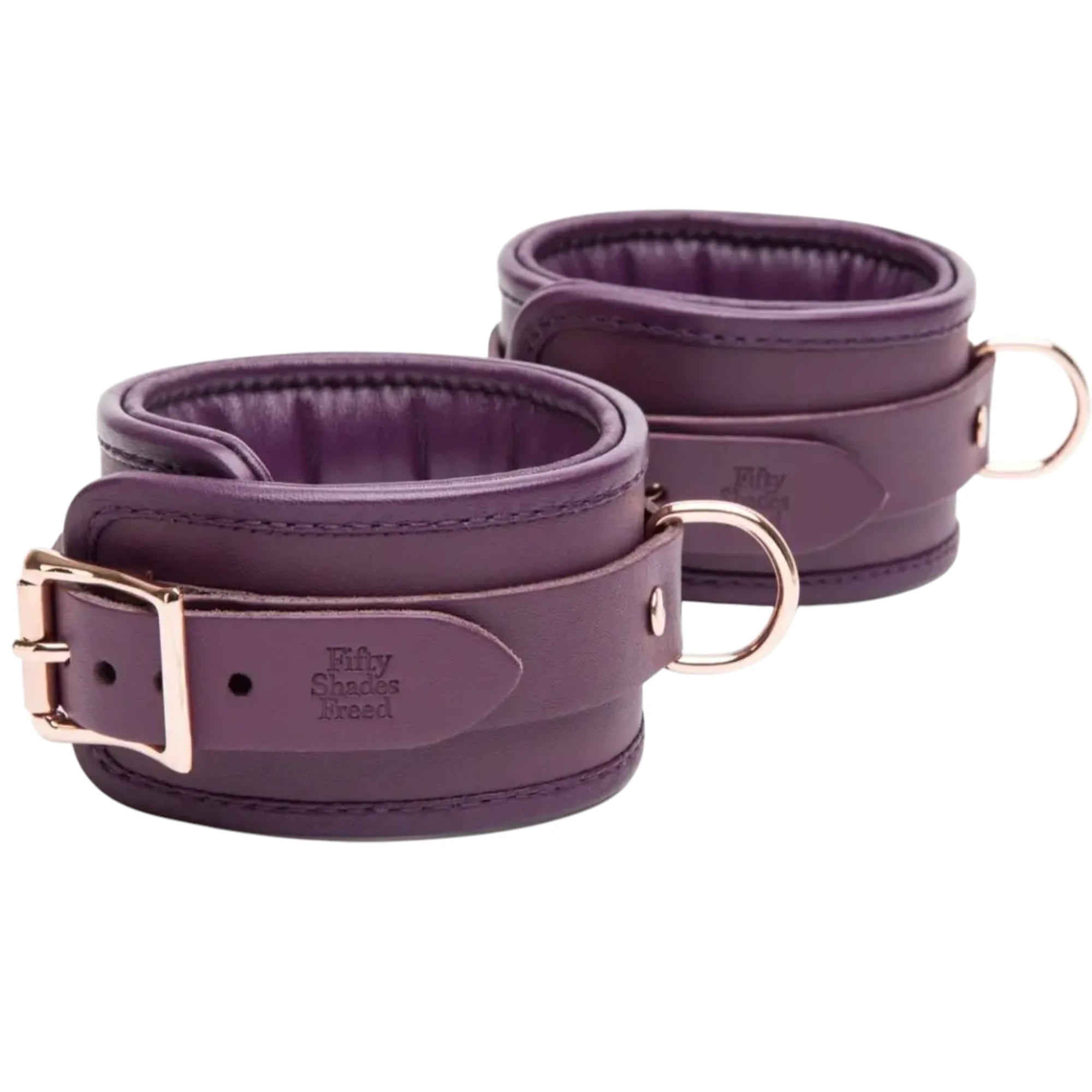 Close-up of the Fifty Shades Freed Cherished Collection leather ankle cuffs in purple, showing the padded lining, embossed Fifty Shades Freed branding, rose-gold buckle hardware and D-ring attachment points against a white background.