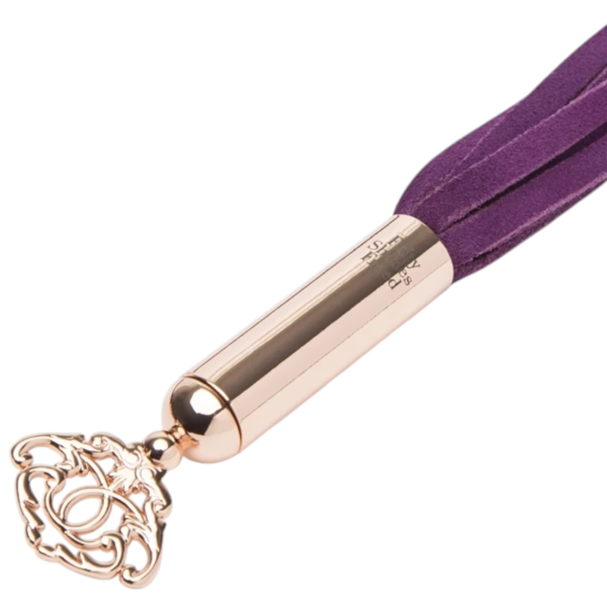Close-up of the Fifty Shades Freed Cherished Collection mini flogger handle, showing the rose-gold metal grip with Fifty Shades Freed branding, the attached purple faux-leather tails and the ornate key-shaped charm at the end against a white background.