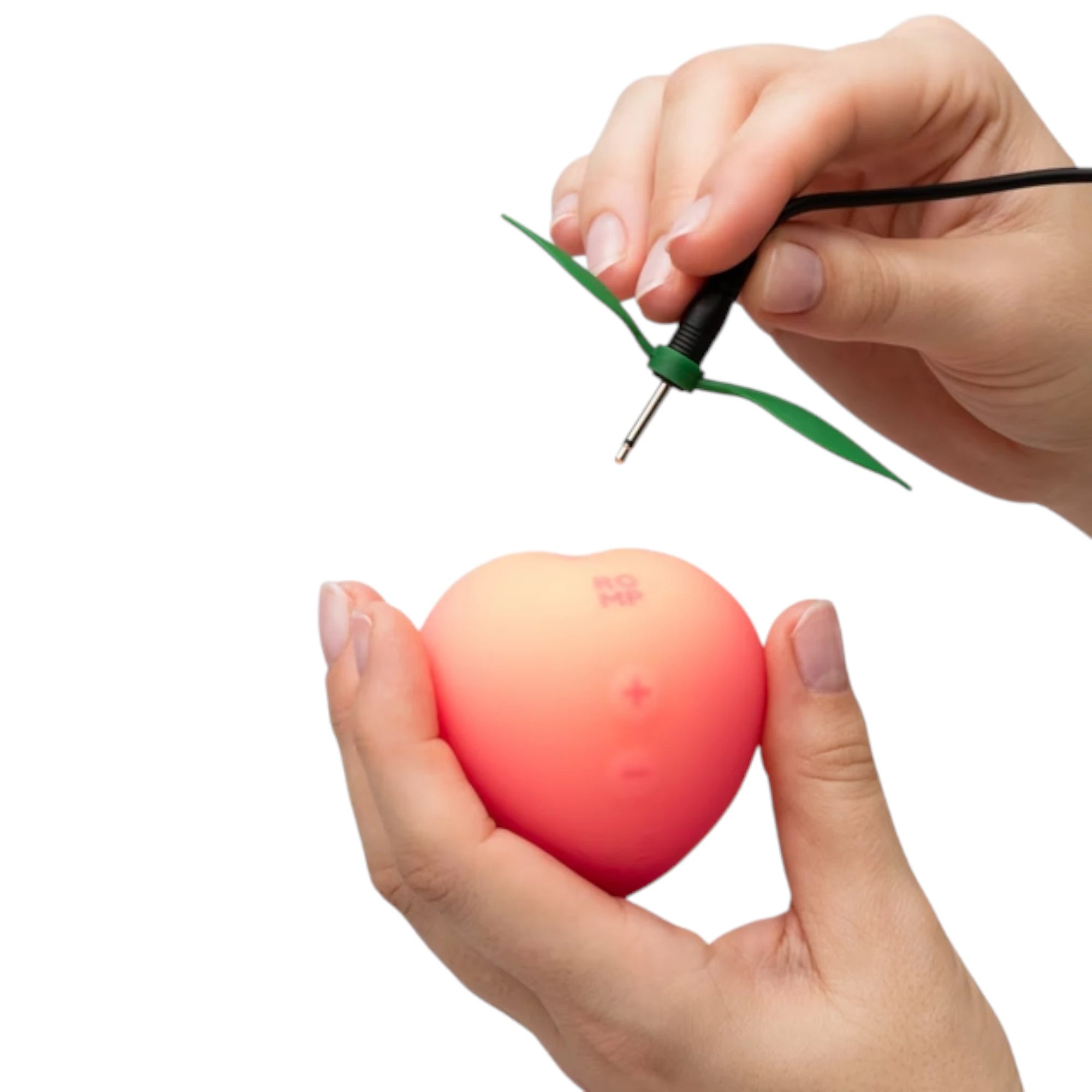 The ROMP Pink Peach rechargeable clitoral stimulator in peach-shaped pink silicone shown held in a hand, with a charging cable and green leaf-shaped charging attachment above it, on a plain white background
