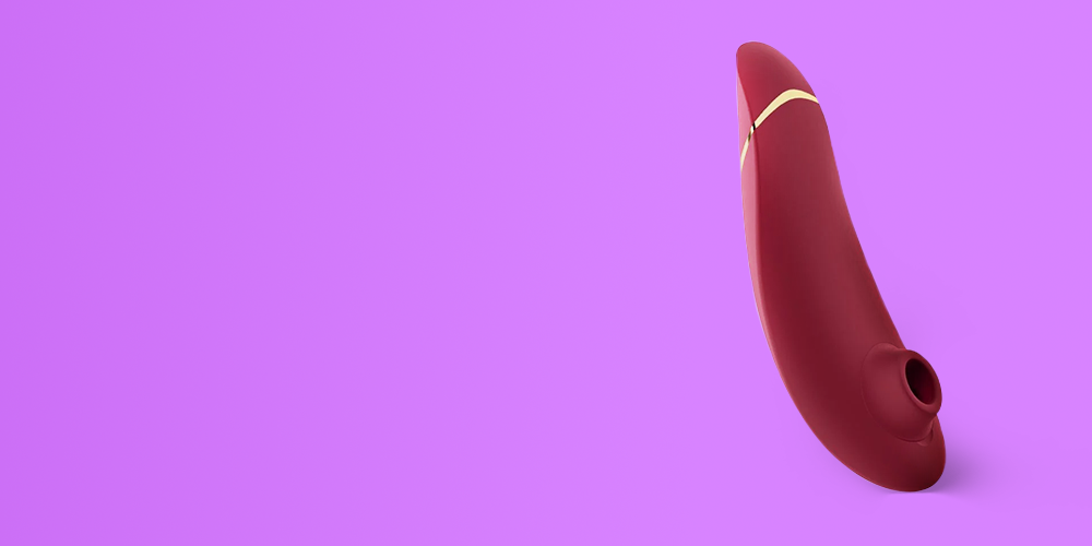 Curved clitoral suction vibrator in deep burgundy silicone with a rounded bulbous head and a gold-toned accent band near the top, positioned vertically on the right side of a vibrant purple gradient background with large empty space on the left