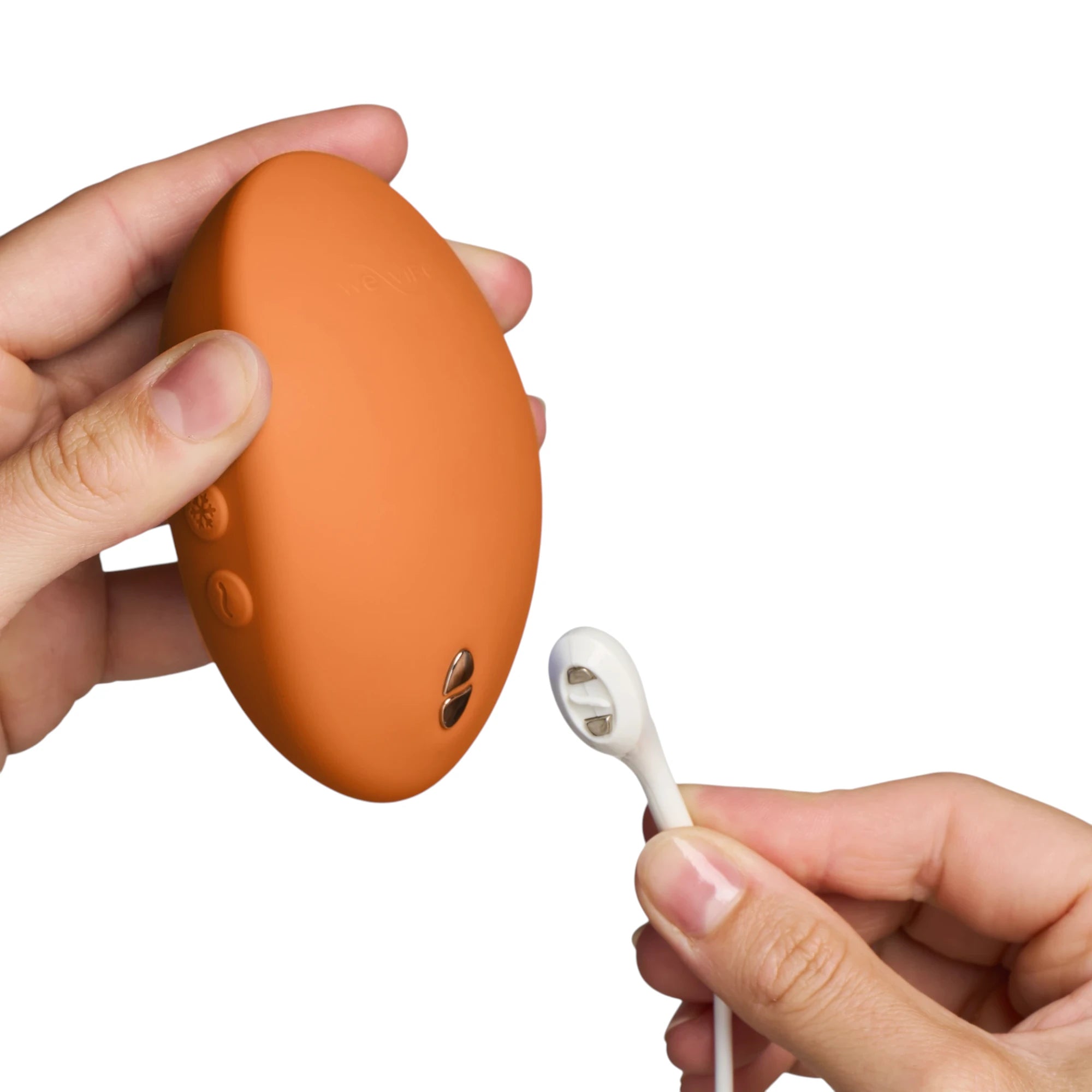 We-Vibe Hot+Cold clitoral vibrator in terracotta orange held in hand displaying oval body, dual motor control buttons, smooth silicone finish, shown with white charging cable against white background